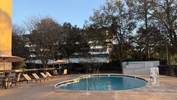 Una piscina con sillas de descanso y sombrillas, rodeada de árboles y un edificio al fondo.