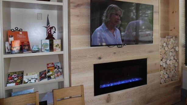 A living room with a flat-screen TV mounted on a wooden wall, a fireplace below, and a bookshelf with various items. - Wingate by Wyndham Jacksonville FL (Jacksonville)