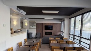 A modern dining area with multiple tables and chairs, a flat-screen TV mounted on the wall, and large windows providing a view of the outdoors. - Wingate by Wyndham Jacksonville FL (Jacksonville)