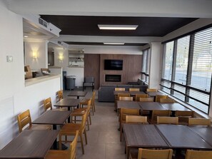 A modern dining area with multiple tables and chairs, a flat-screen TV mounted on the wall, and large windows providing a view of the outdoors. - Wingate by Wyndham Jacksonville FL (Jacksonville)