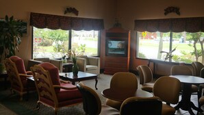 Ein Zimmer mit Fernseher, mehreren Stühlen, einem Tisch und großen Fenstern mit Blick auf Grünflächen.