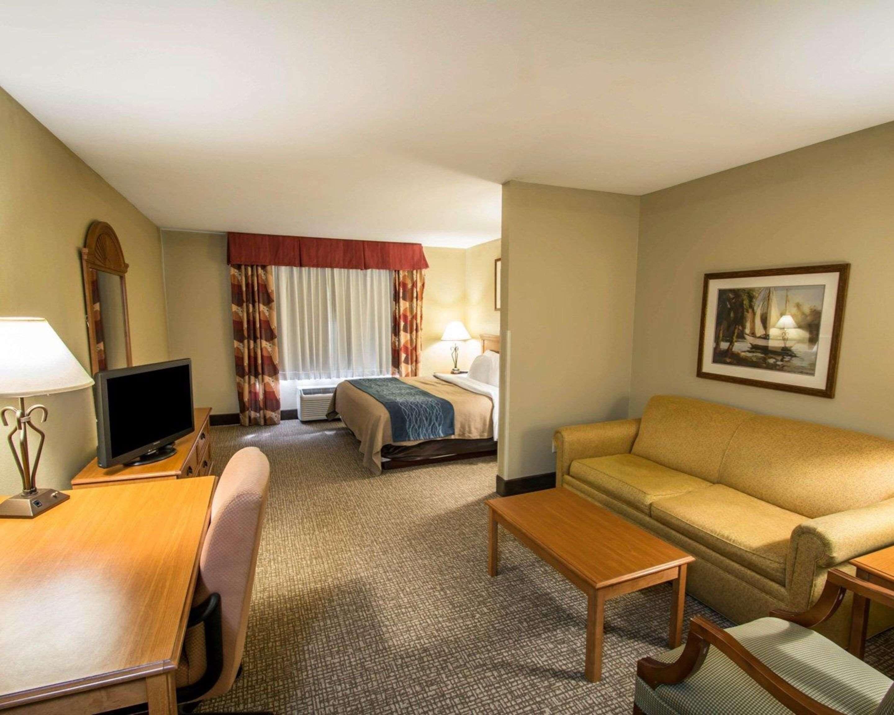 A hotel room with a bed, a sofa, a desk with a computer, a chair, a lamp, and a painting on the wall.