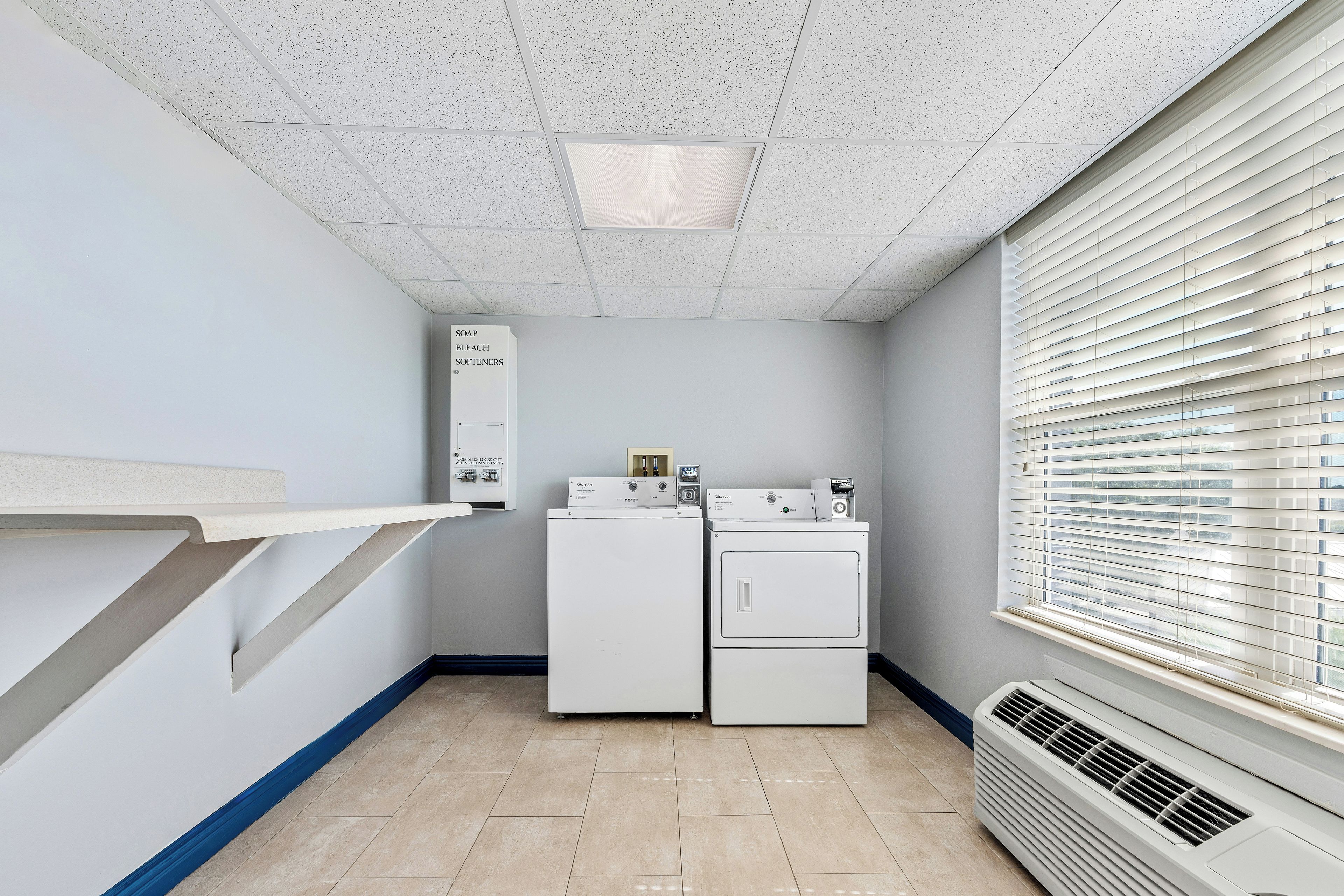 A laundry room with a washer and dryer, a shelf with cleaning supplies, and a window with blinds.