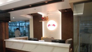 A modern reception area with a marble counter, a large illuminated sign, and a view of the outside through large windows. - Manyavar Guest House (Deoghar)