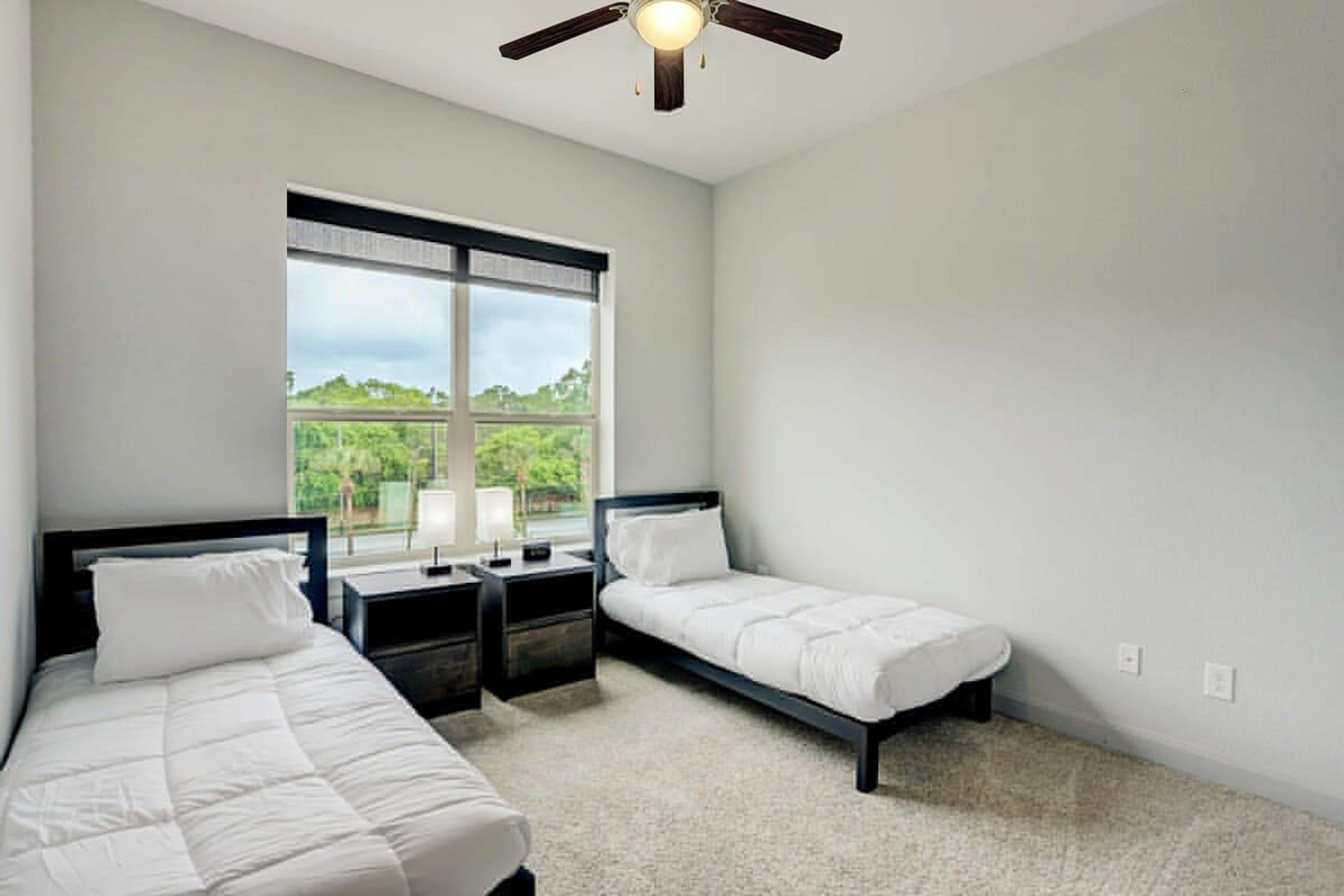 A bedroom with two single beds, a window with a view of greenery, a ceiling fan, and a nightstand with a lamp.