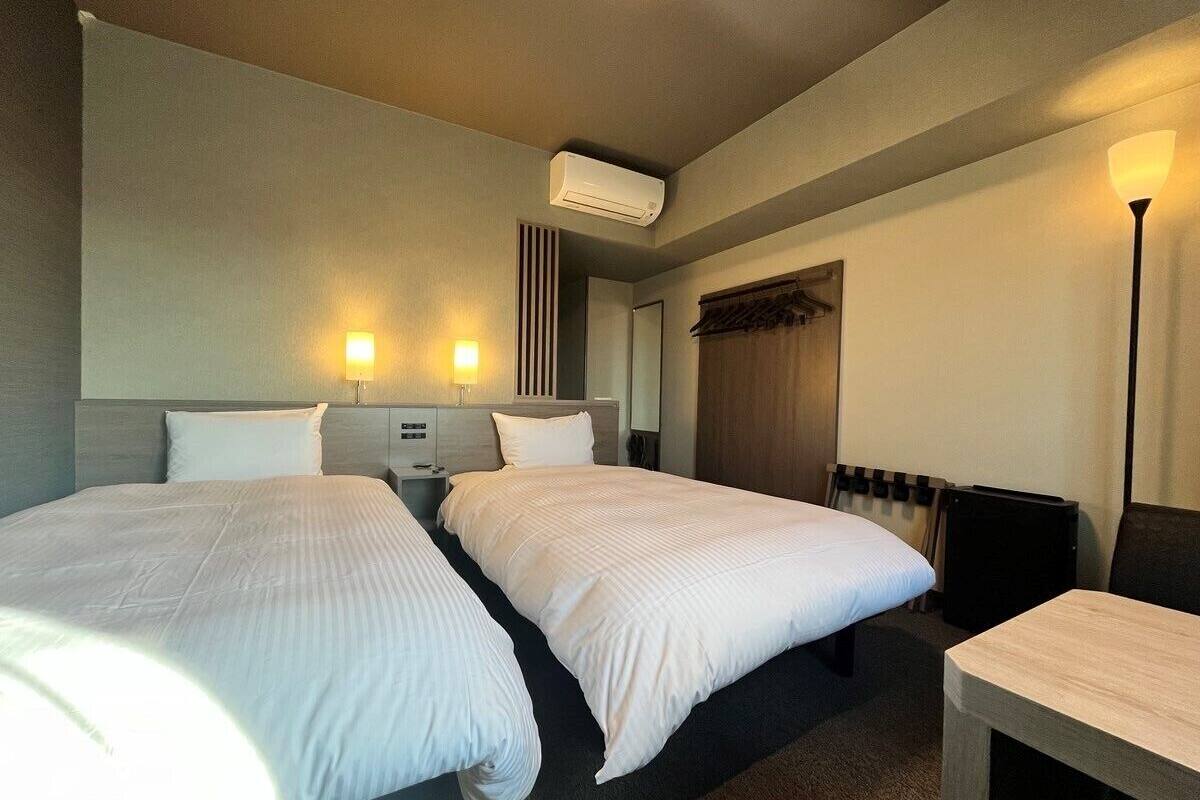 A hotel room with two beds, a desk, a lamp, an air conditioner, and a radiator.