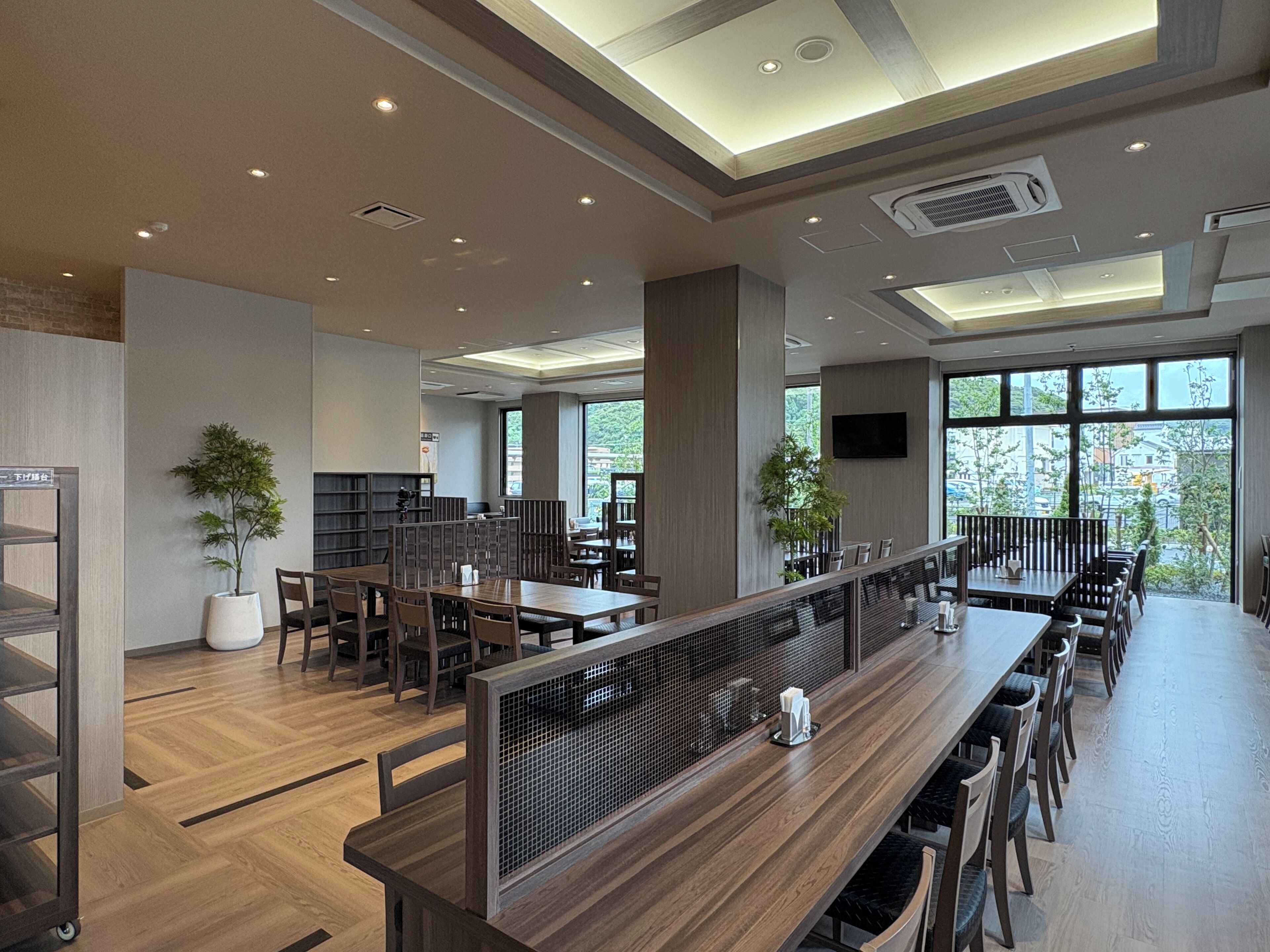 Un comedor moderno con mesas y sillas de madera, un ventanal que ofrece vistas al exterior e iluminación empotrada en el techo.