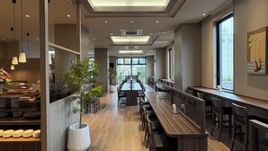 Un comedor moderno con largas mesas y sillas de madera, una planta grande en maceta y vistas a través de las ventanas.