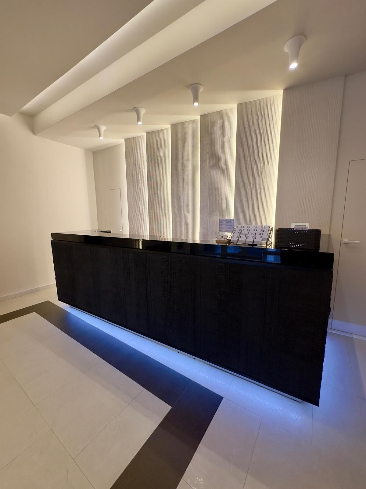 A modern reception desk with a black surface and illuminated edge, flanked by vertical light panels, in a minimalist interior with white walls and a tiled floor.