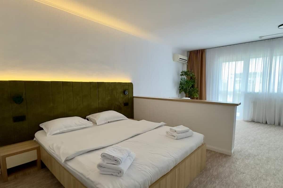 Ett hotellrum med en stor säng, ett nattduksbord, ett litet skrivbord, en växt och ett fönster med gardiner.