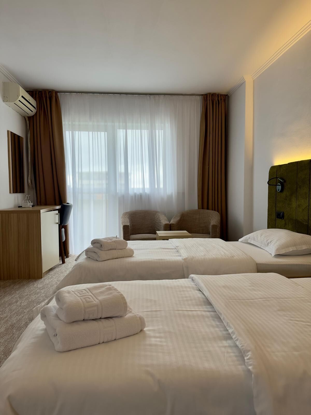 A hotel room with two beds, a desk, a chair, and a large window with curtains.
