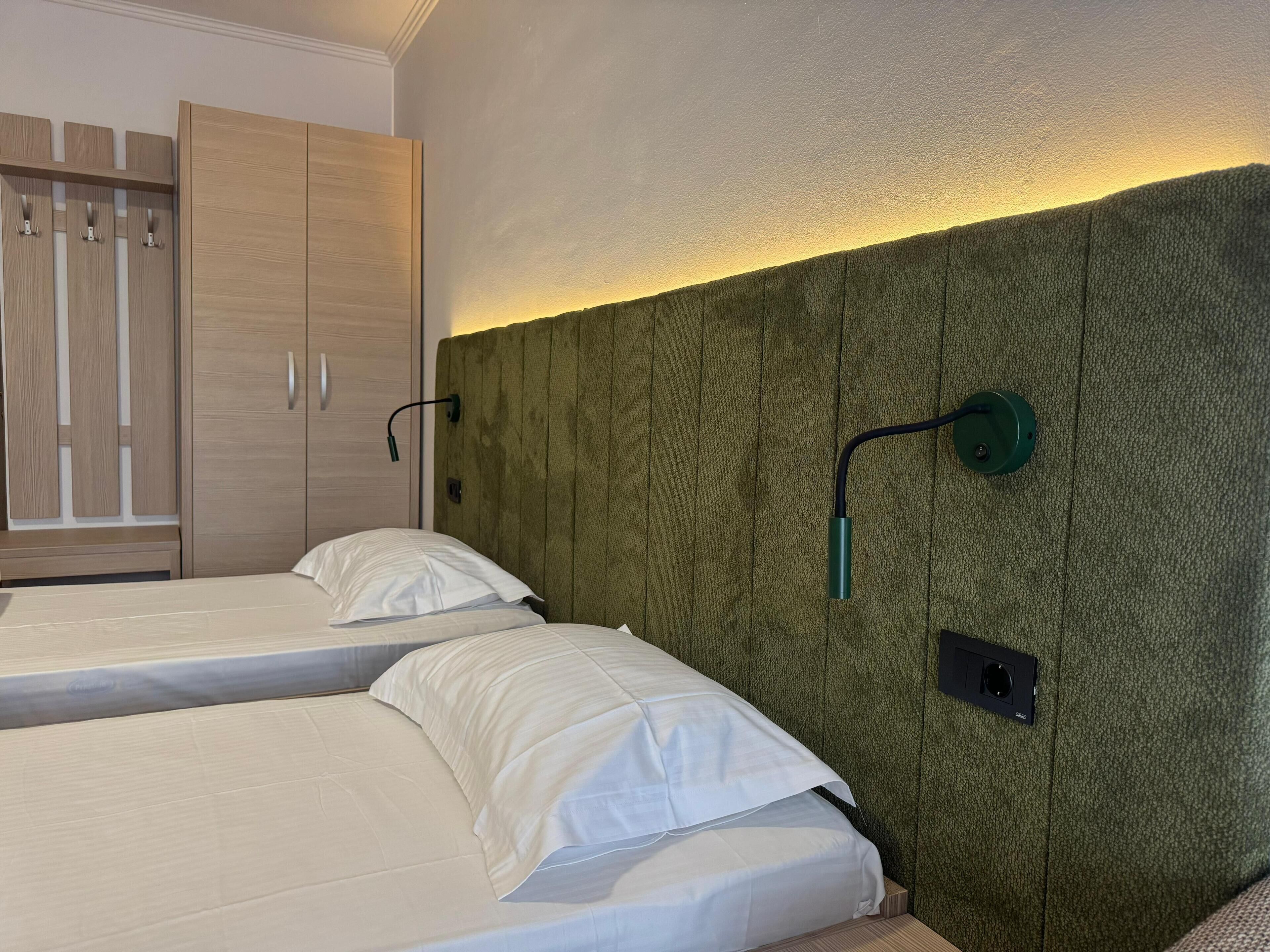 A bedroom with two single beds, a wooden wardrobe, and a green textured headboard with wall-mounted lighting.