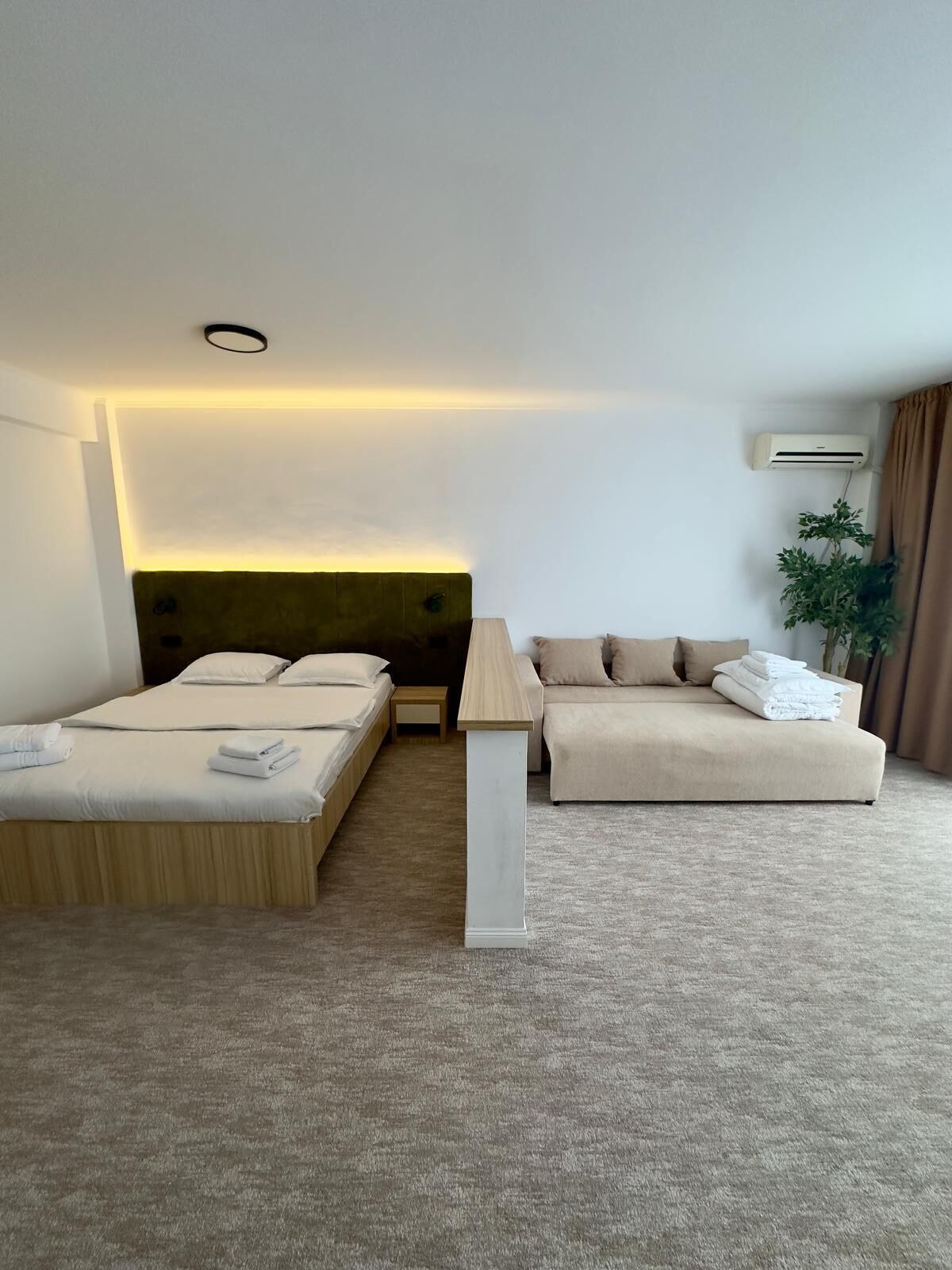 A hotel room with a bed, a sofa, a bedside table, and an air conditioning unit.