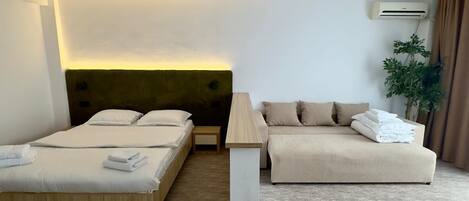 A hotel room with a bed, a sofa, a bedside table, and an air conditioning unit.