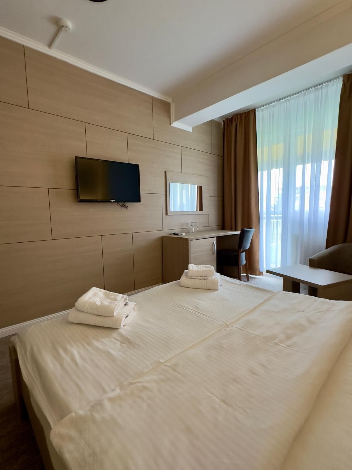 Une chambre d’hôtel avec un grand lit, un téléviseur fixé au mur, un bureau avec une chaise et une fenêtre avec des rideaux.