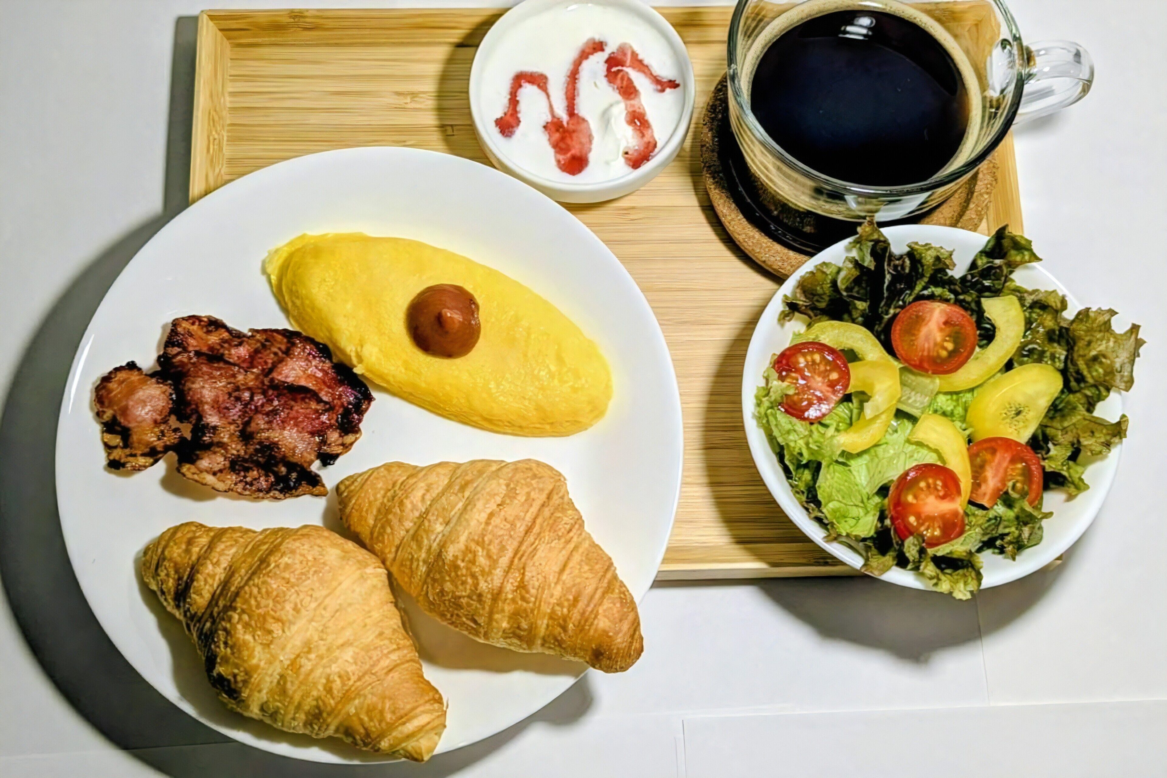 Un plato de desayuno con cruasanes, tocino y huevos revueltos, un tazón de ensalada, una taza de café y un vasito de yogur con trocitos de tocino.