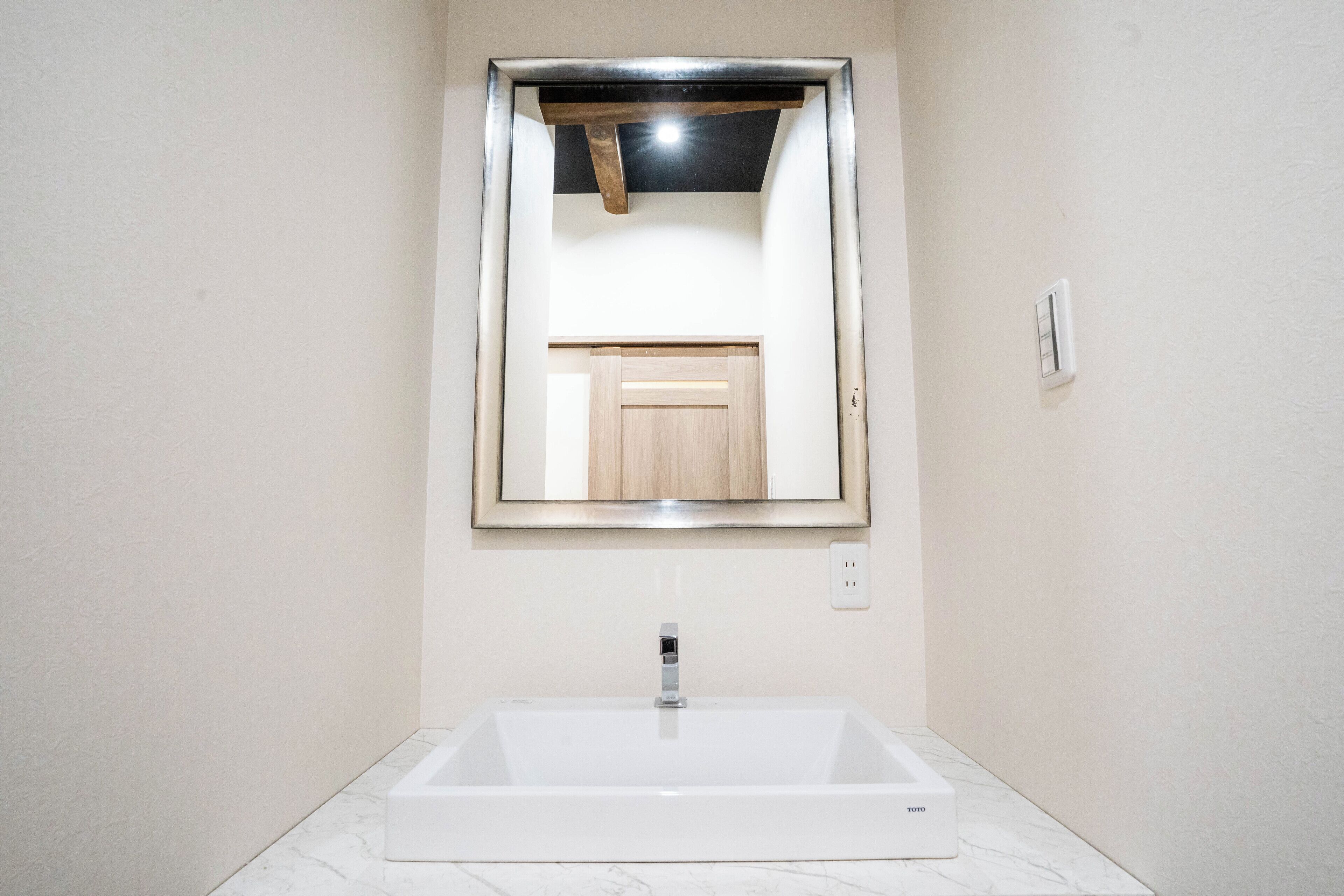 Un baño con un lavamanos blanco rectangular, un espejo con marco de madera y un interruptor de luz en la pared.