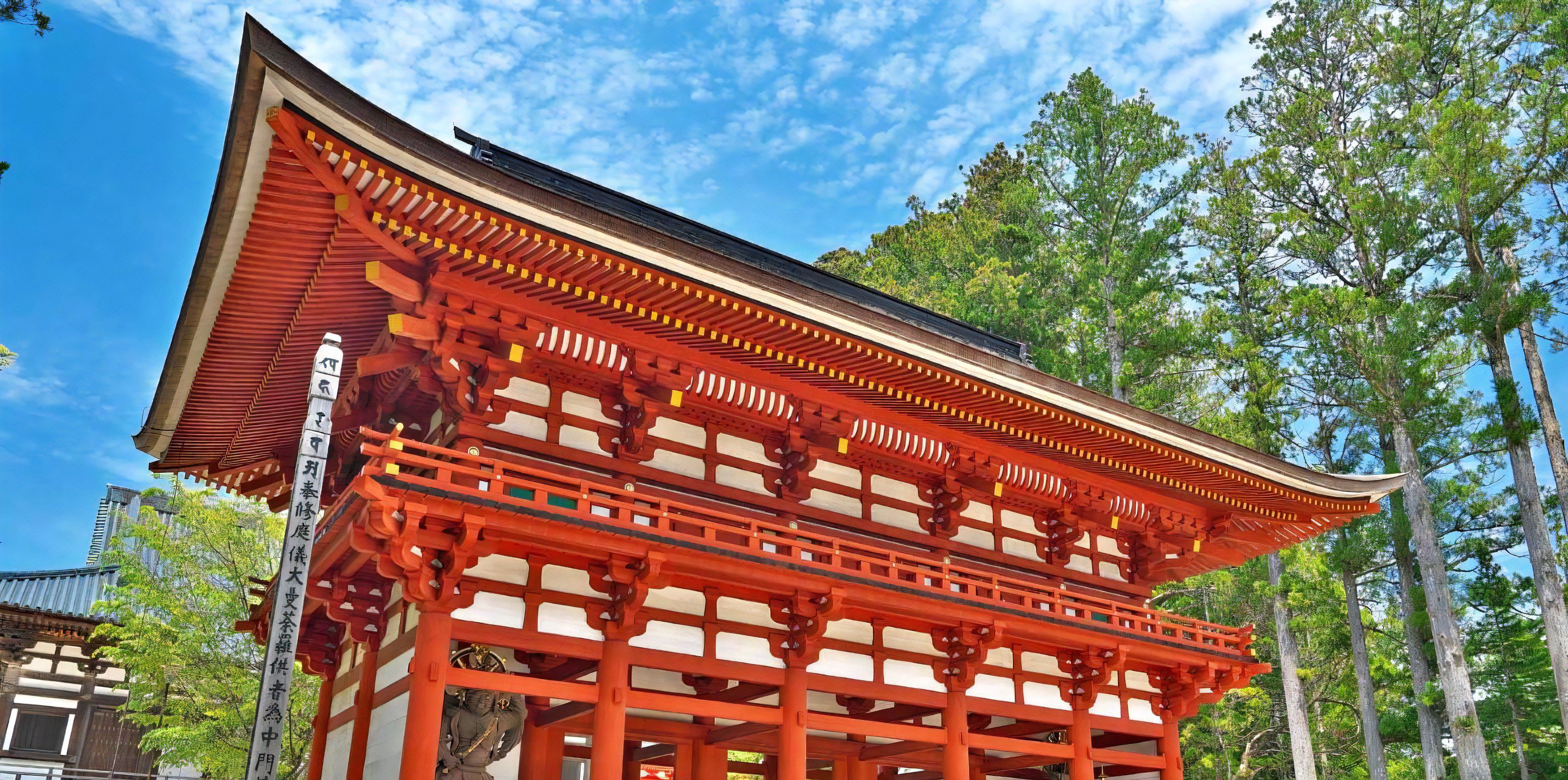 Un portail japonès tradicional amb una combinació de colors vermell i blanc, davant d'un fons de paisatge arborat i cel blau.