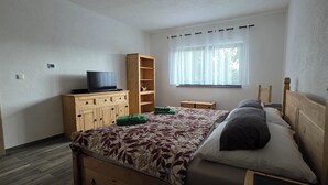A bedroom with a bed, wooden wardrobe, a television on a stand, a window with sheer curtains, and a wooden chair.