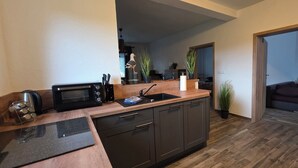 A compact kitchen with a microwave, toaster, and sink. There are cabinets and a countertop with a potted plant. A doorway leads to a living area with a couch and a plant.