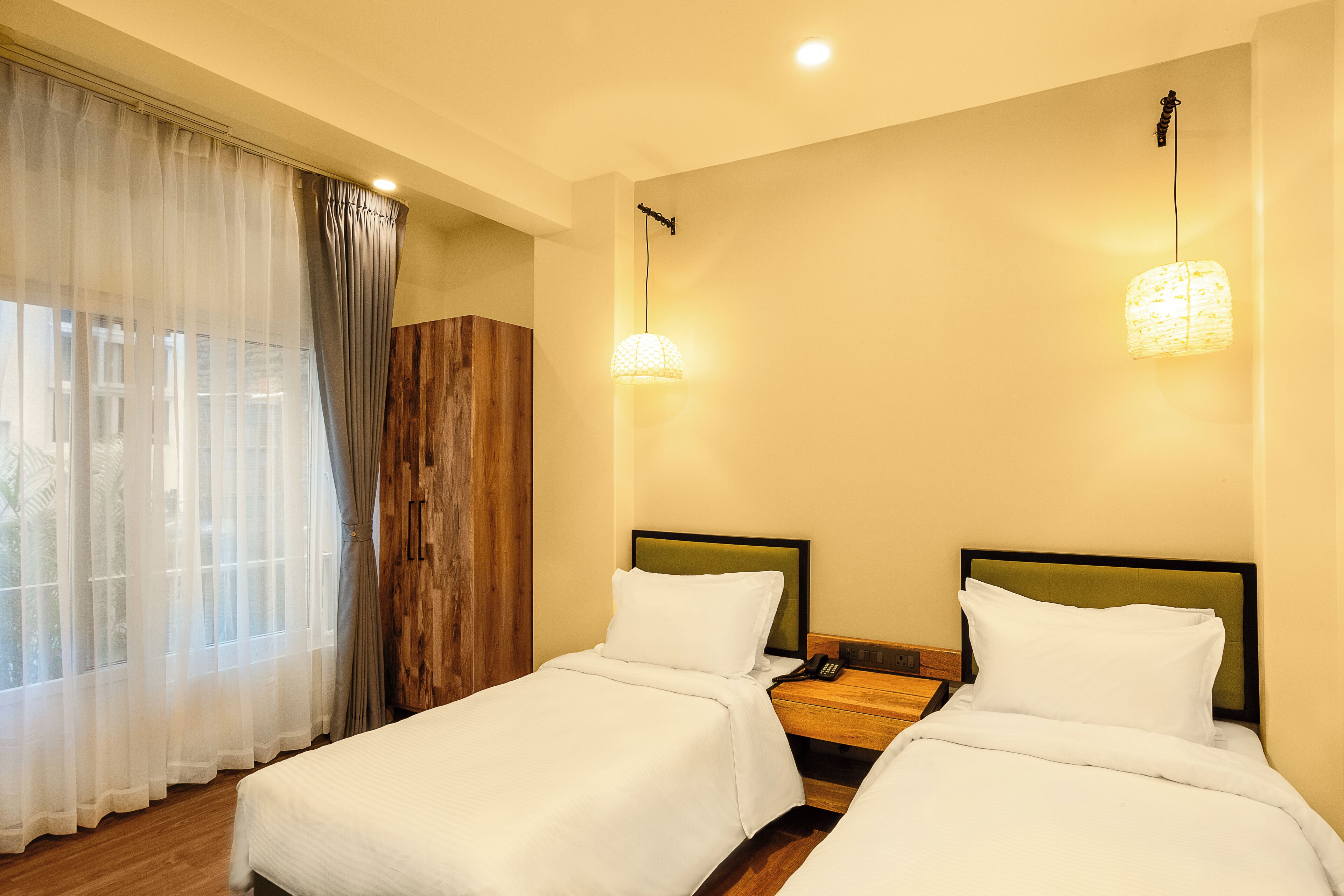A hotel room with two beds, a wooden wardrobe, a nightstand with a lamp, and sheer curtains.