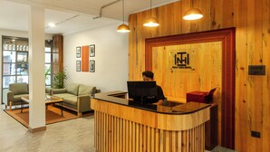 Un comptoir d’accueil avec un revêtement en bois, un ordinateur et un téléphone rouge. Des tableaux encadrés ornent le mur et une plante est placée dans un coin.