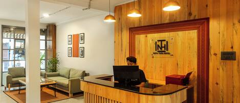 A reception desk with a wooden finish, a computer, and a red telephone. There are framed pictures on the wall and a plant in the corner.