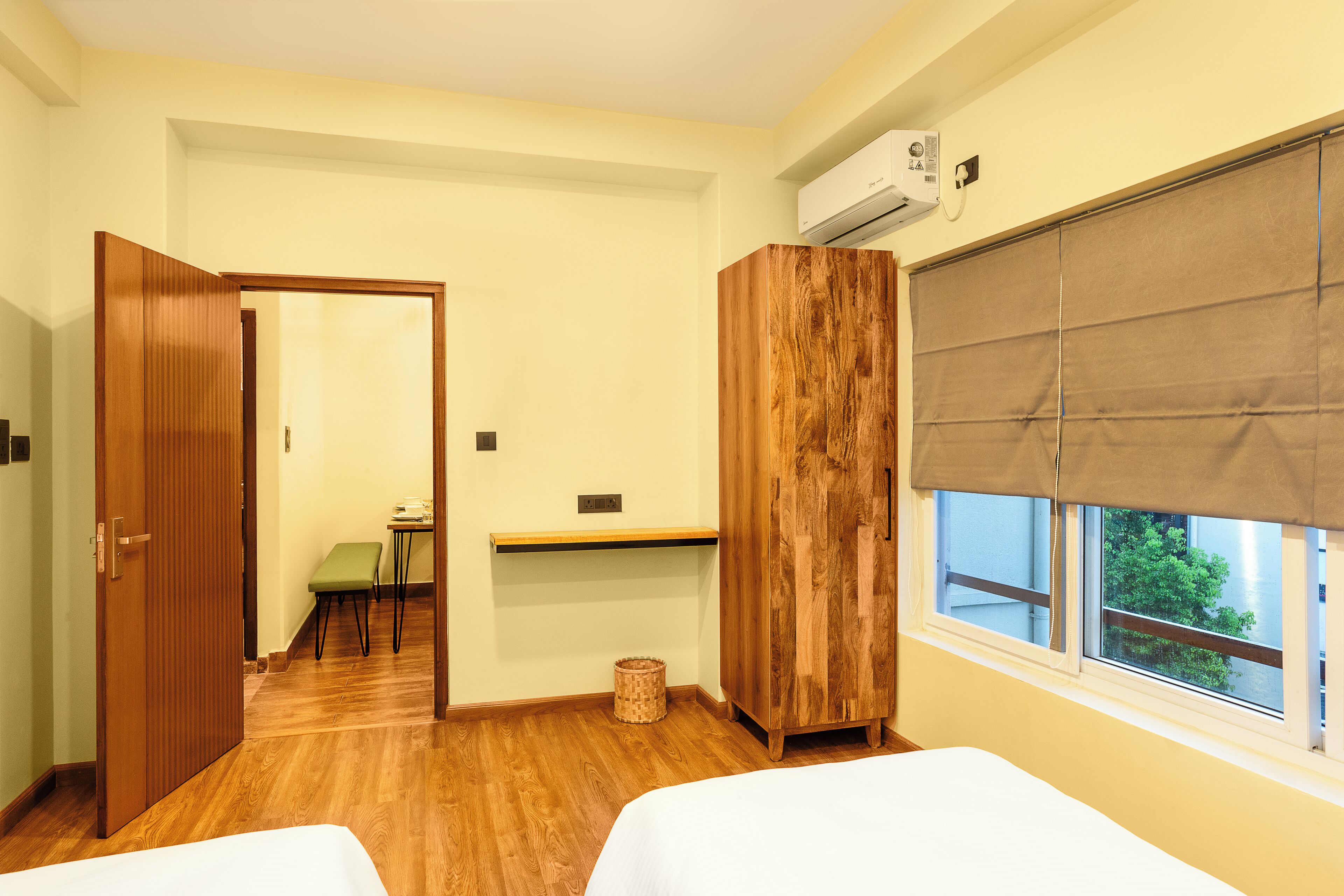 Une chambre d’hôtel avec deux lits, une armoire en bois, une petite table avec un banc, une fenêtre donnant sur un paysage agréable et un appareil de climatisation.