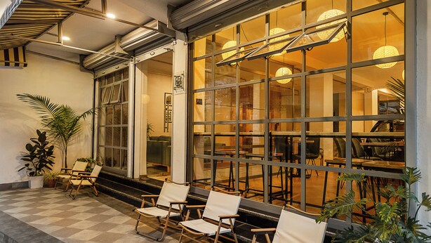 L’entrée d’un hôtel avec un sol à carreaux, un espace extérieur aménagé avec des chaises, et de grandes fenêtres éclairées de l’intérieur.