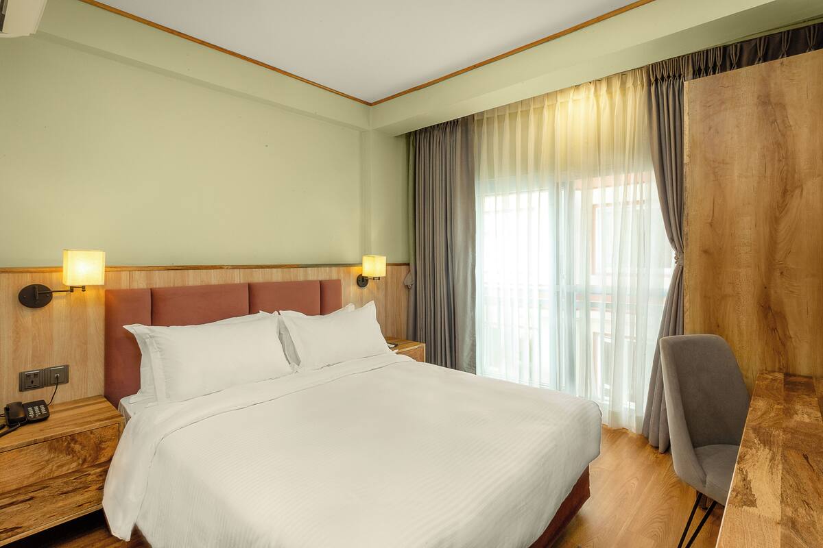Camera d'albergo con un letto grande, una testata in legno, due lampade da comodino, un comodino in legno con un telefono e una sedia.