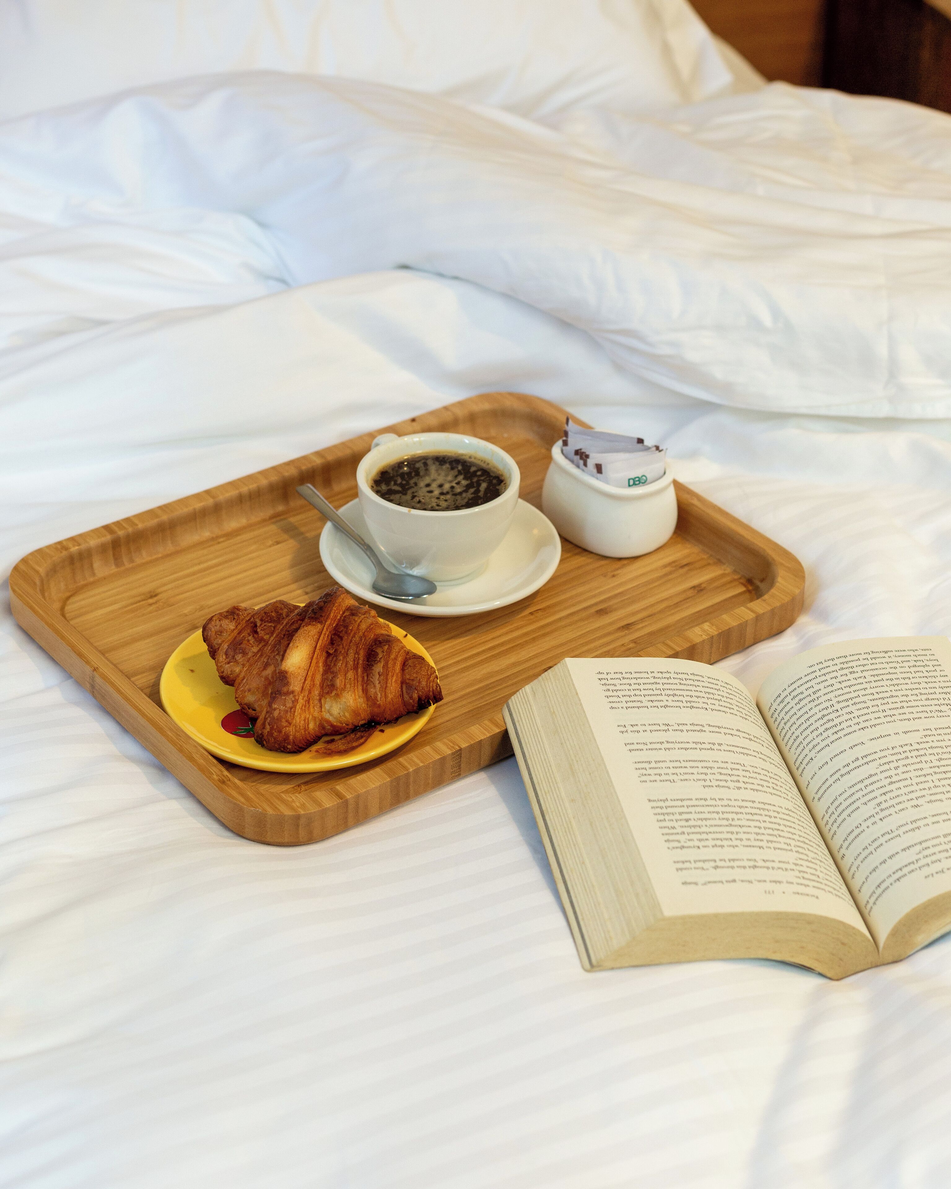 A wooden tray with a croissant, a cup of coffee, and a small container of cream on a bed with white sheets.