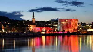 A waterfront cityscape with illuminated buildings, a church steeple, and a calm river reflecting the lights. - Apartment in Linz Near Danube Promenade (Linz)