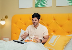 A person sitting on a bed with a yellow headboard, eating a meal and drinking from a glass.