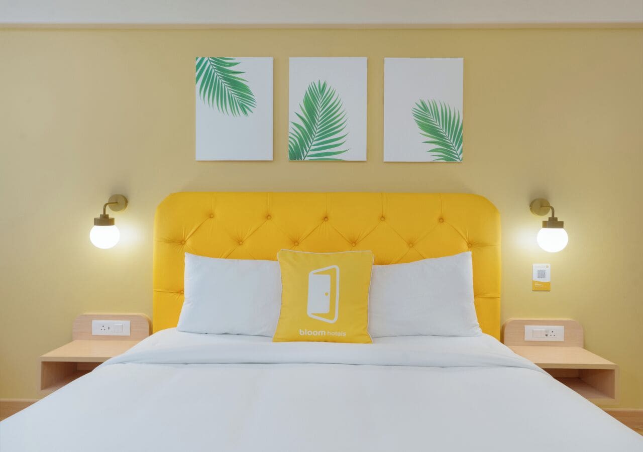 A hotel room with a yellow headboard, white bedding, and three palm leaf wall art pieces.