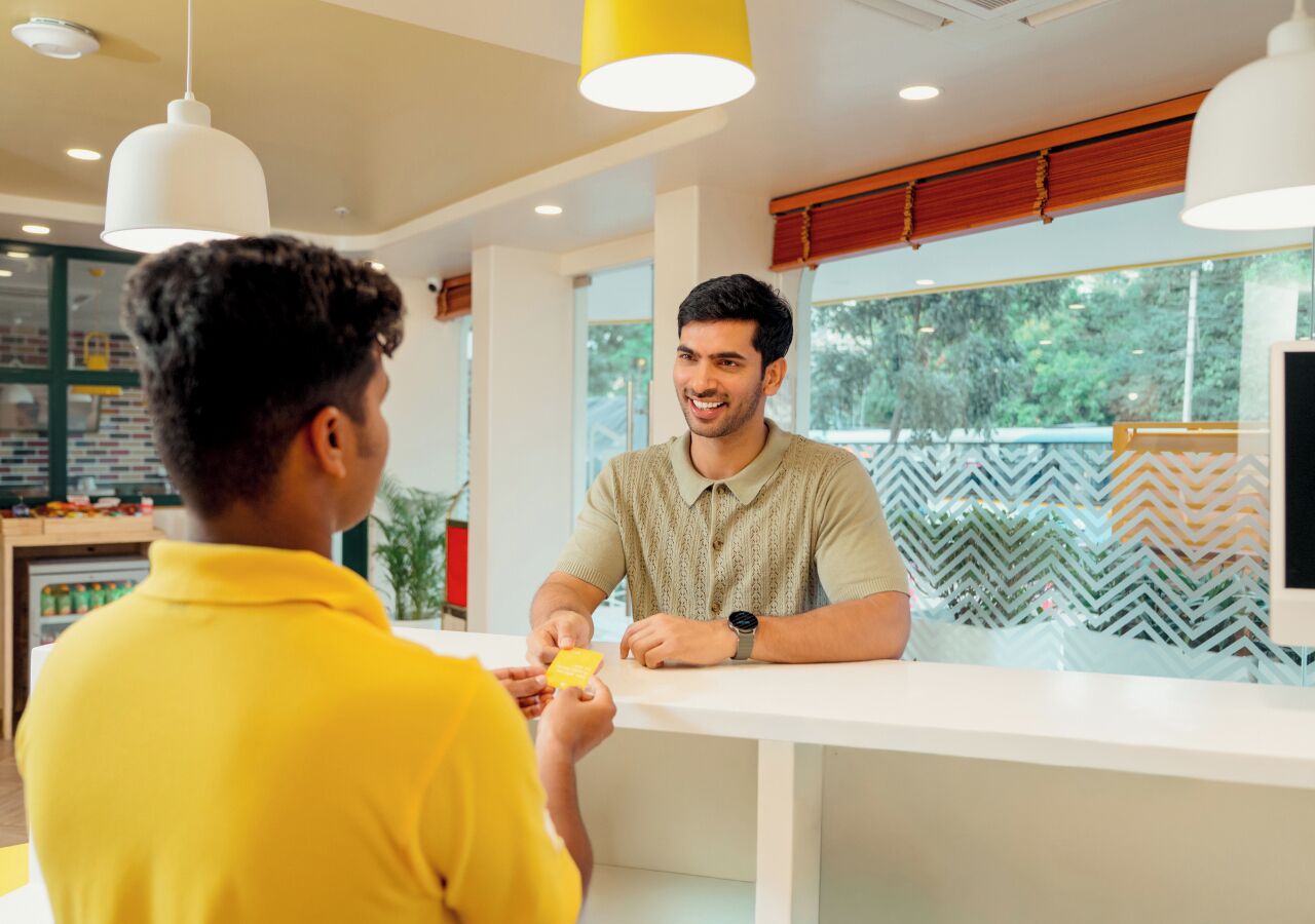 Una persona con una camicia gialla interagisce con un bancone in un ambiente interno luminoso, con ampie finestre e lampade a sospensione moderne.