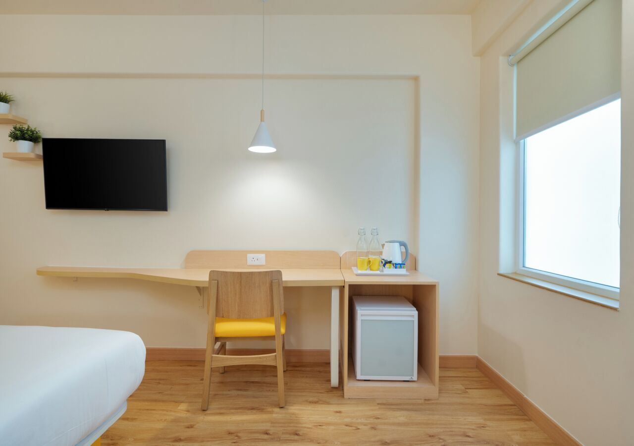 Une chambre comprenant un lit, un bureau avec une chaise, un petit réfrigérateur, un téléviseur fixé au mur et une fenêtre laissant entrer la lumière naturelle.