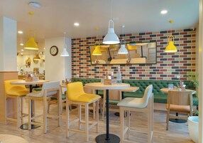 A modern restaurant with round tables and yellow chairs, brick-patterned walls, and hanging pendant lights.