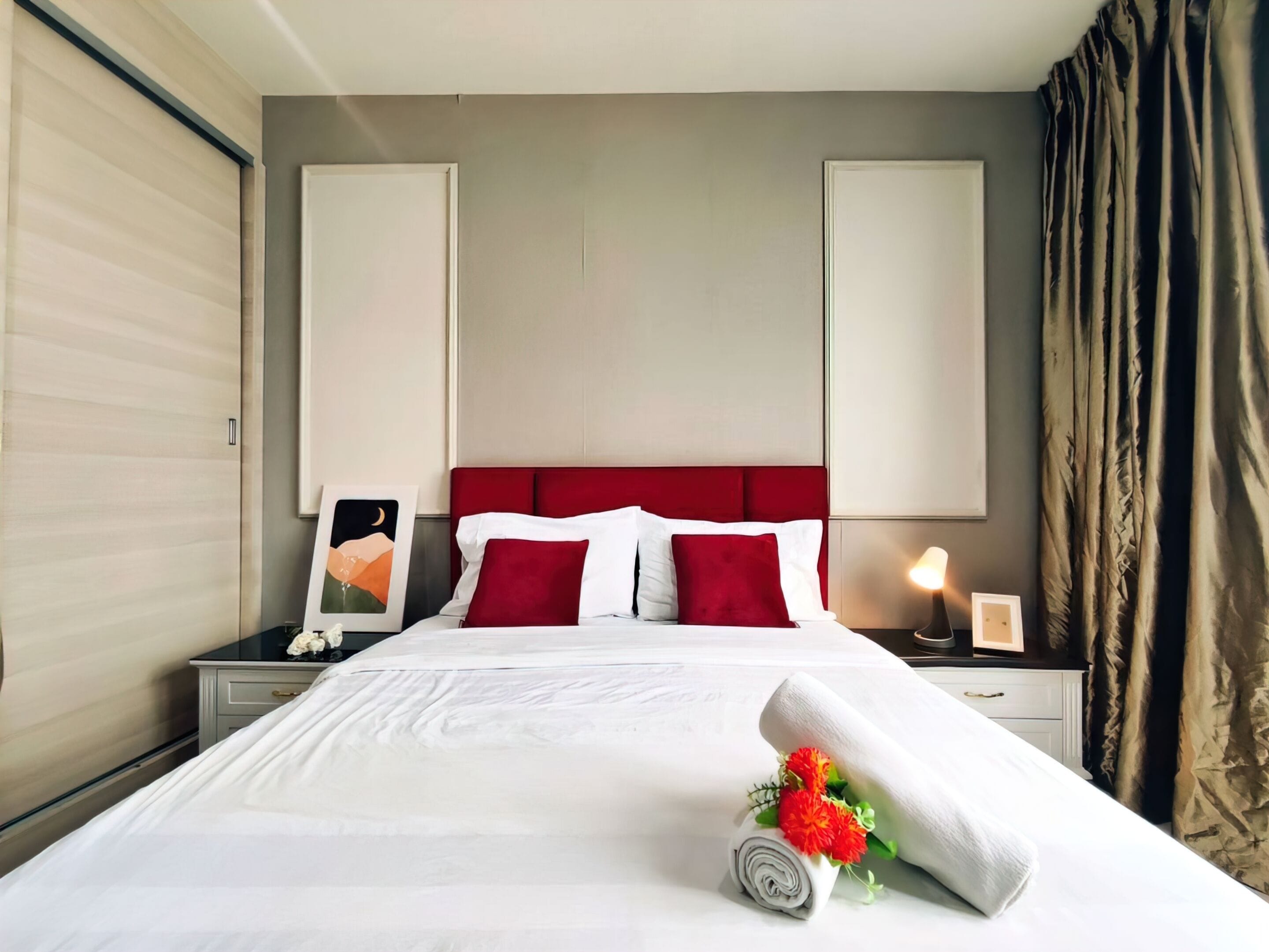 A hotel room with a neatly made bed, a red headboard, and a nightstand with a lamp and framed pictures.