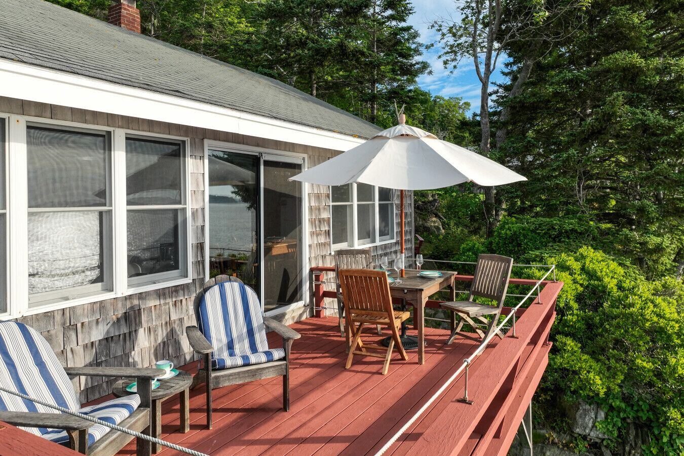 A wooden deck with a table and chairs, a large umbrella, and a view of a forested area.
