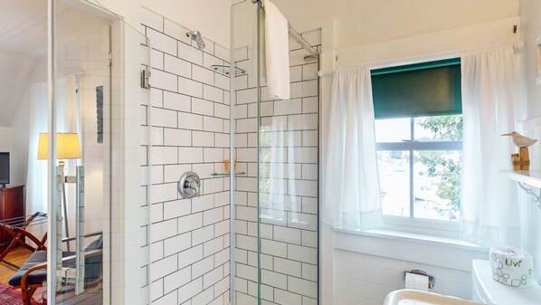 A bathroom with white tiled walls, a glass shower enclosure, a window with a green blind, and a toilet paper dispenser.
