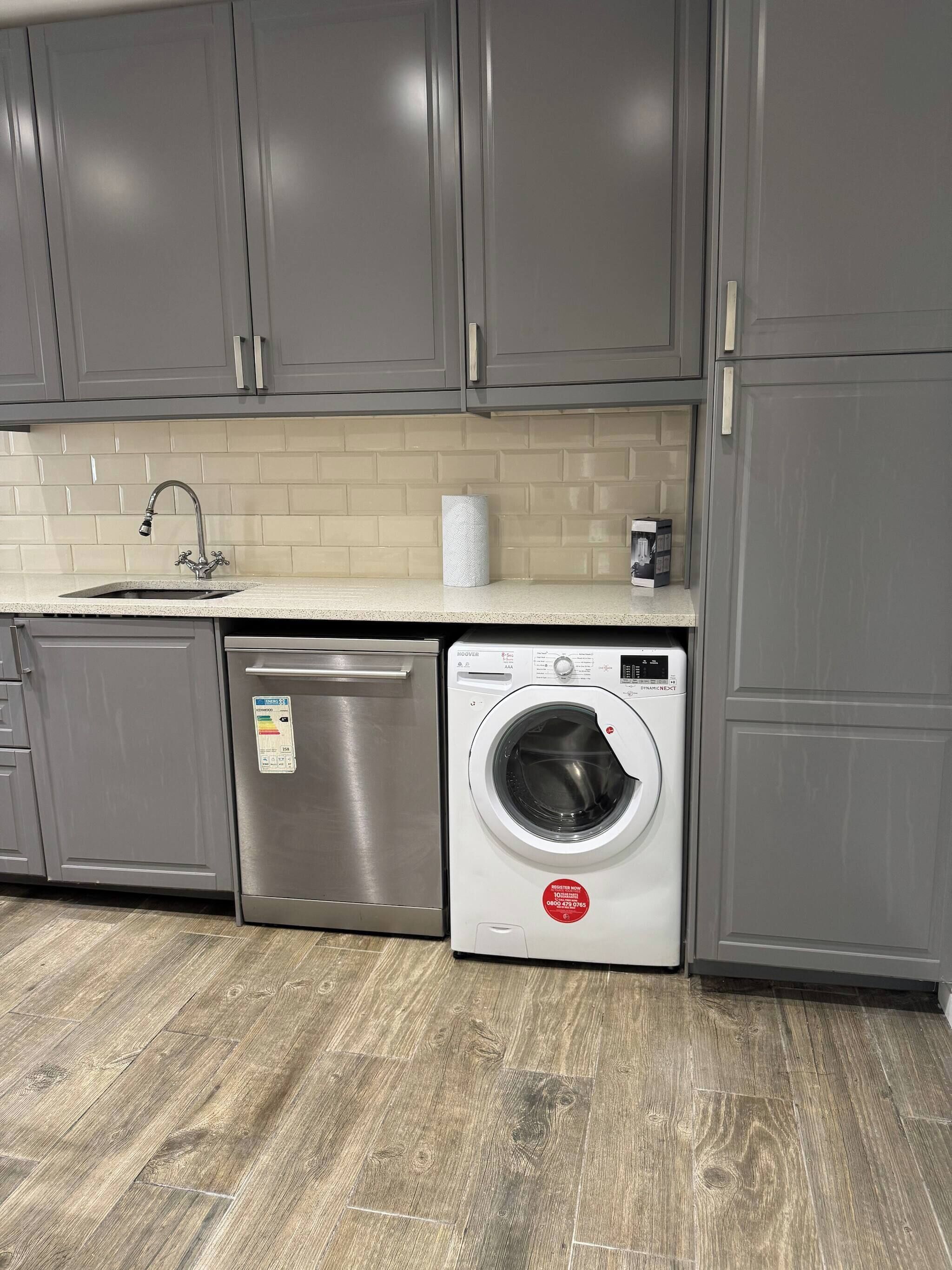 A kitchen with grey cabinets, a stainless steel dishwasher, a washing machine, and a tiled backsplash.
