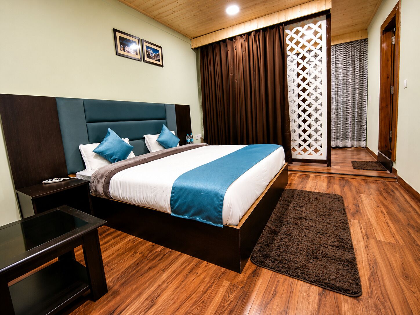 A modern hotel room with a large bed, wooden flooring, a nightstand, and a view of a balcony with curtains.
