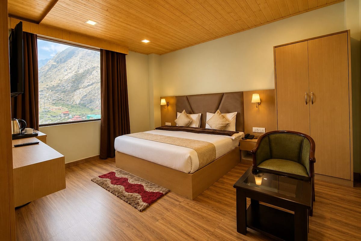 A hotel room with a bed, bedside table, chair, wardrobe, and a view of mountains.