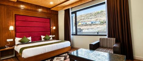 Habitación de hotel con una cama grande, una mesa de centro de vidrio, un sillón a rayas y vistas a un paisaje desértico.