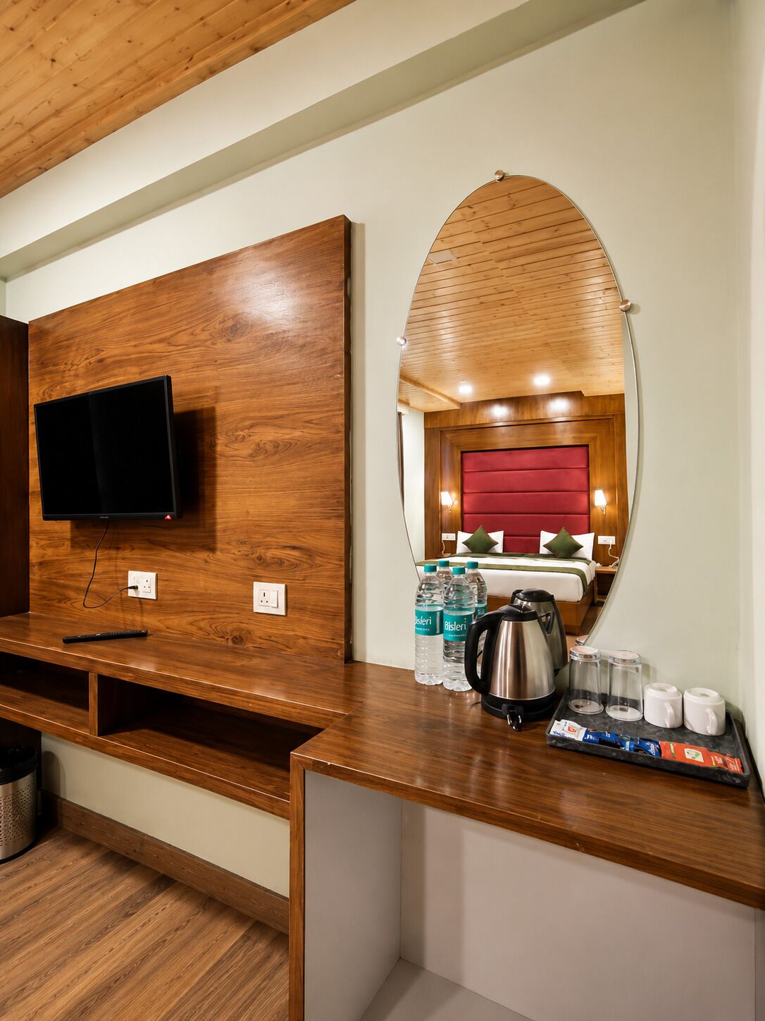 A hotel room with a wooden vanity, a mounted television, a mirror, and a kitchenette area with a kettle and water bottles.