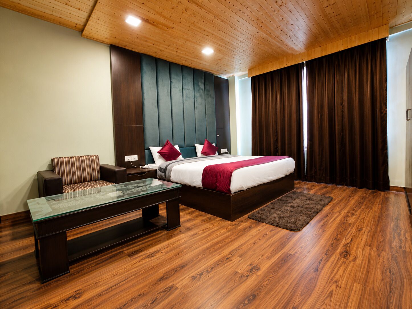 A modern bedroom with a wooden ceiling, a large bed with red bedding, a dark headboard, a glass coffee table, and a brown rug.