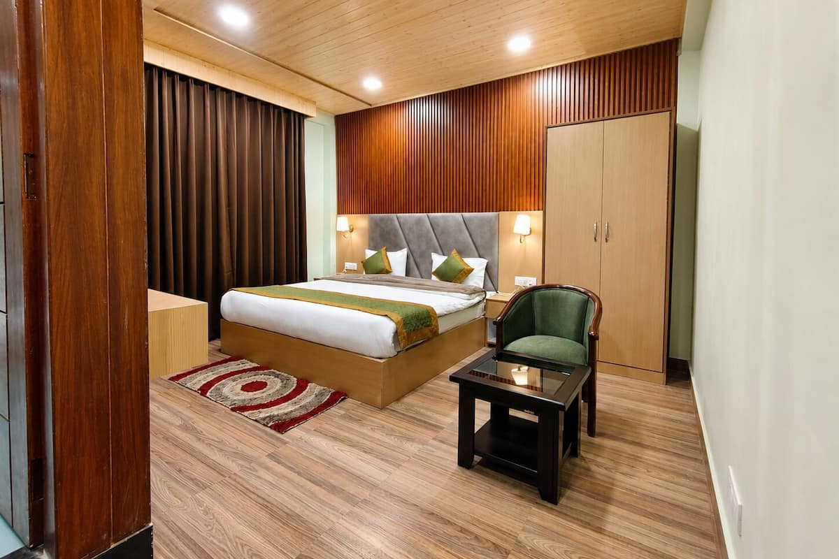 A modern hotel room with a large bed, a wooden headboard, a green armchair, a small side table, and a wardrobe.