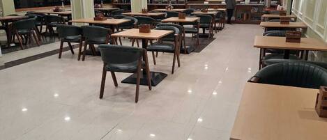 A spacious dining area with multiple tables and chairs, a kitchen area in the background, and a polished tile floor.