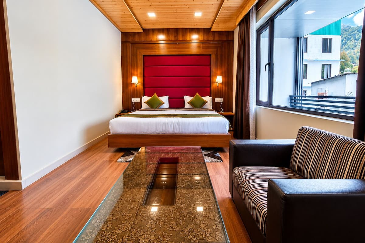 A modern hotel room with a large bed, a sofa, a glass coffee table, and a view of the outside.