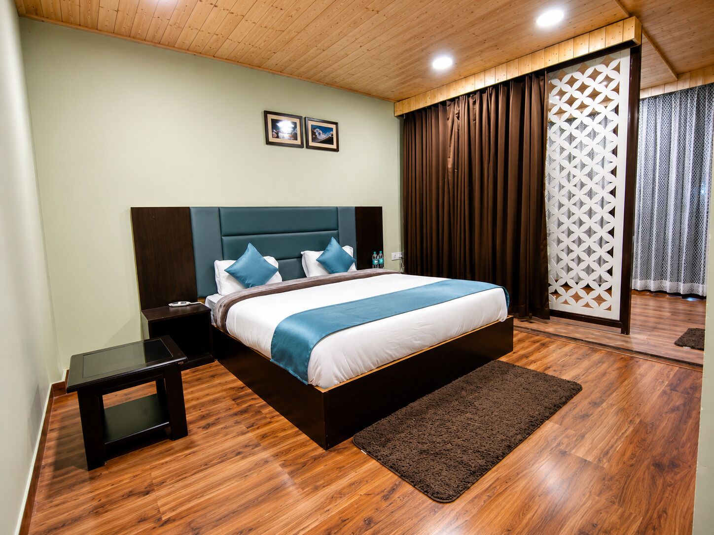 A bedroom with a bed, bedside tables, a wooden floor, a brown rug, and a ceiling with wooden beams and recessed lighting.